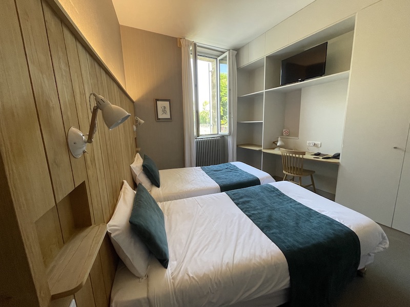 Chambre de l'Auberge de la Commanderie à Saint-Emilion. Une grande tête de lit en bois sur la gauche, un grand bureau encadré d'étagères sur la droite rendent cette chambre fonctionnelle et cosy à souhait.