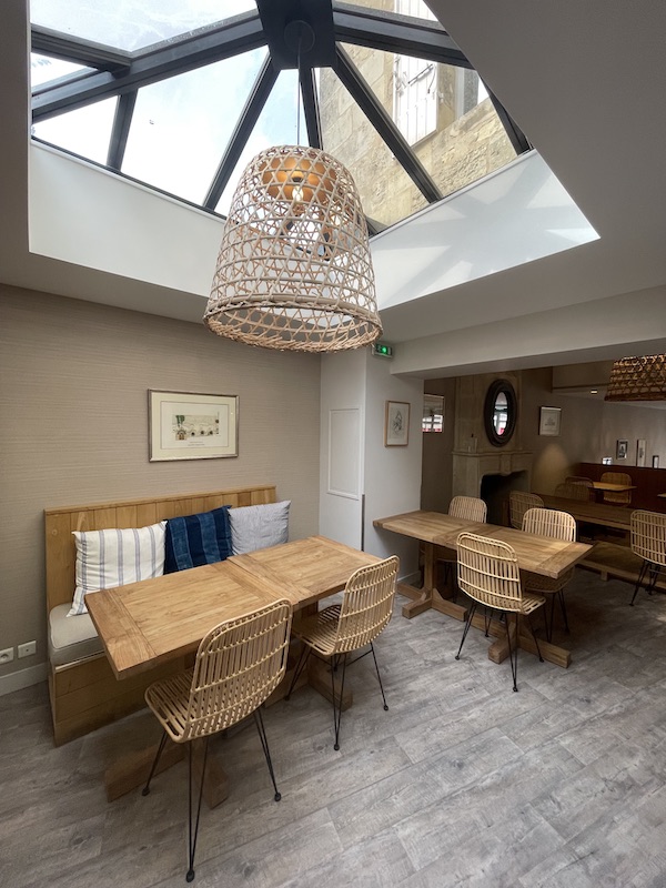 La salle du petit-déjeuner de l'Auberge de la Commanderie à Saint-Emilion mêle tables en bois et chaises en rotin pour une ambiance chaleureuse. Un puit de lumière diffuse la lumière du jour dans toute la pièce.